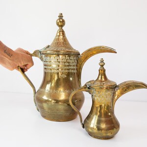 Könnte beinhalten: Zwei antike Kaffeekannen aus Messing mit aufwendigen Details. Die größere Kanne hat einen hohen, konischen Deckel und einen gebogenen Griff. Die kleinere Kanne hat ein ähnliches Design. Beide haben einen warmen, goldenen Farbton und sind vor weißem Hintergrund.