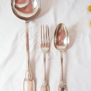 Peut inclure: Ensemble de couverts en argent comprenant une louche, une fourchette et une cuillère. Les ustensiles présentent des détails ornementaux sur les manches et sont disposés sur une nappe blanche. Le bol de la louche reflète l'environnement.