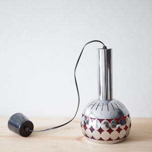 Puede incluir: Lámpara colgante cromada con parte superior cilíndrica y base esférica. La base presenta un patrón rojo en forma de diamante visible a través de recortes. Un cable negro y una pieza cilíndrica negra se extienden desde la lámpara.