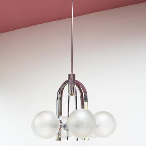 Op de afbeelding: Een chromen kroonluchter met vijf glazen bolvormige lampenkappen. De armatuur heeft een strak, modern ontwerp met een centrale stang en gebogen armen. Het plafond is stoffig roze.