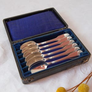 May include: A set of six silver spoons presented in a black box with a blue velvet interior. The spoons feature a decorative handle design and are neatly arranged. The box is open, showcasing the spoons.