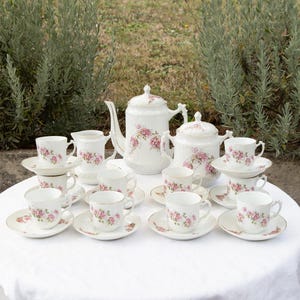 Pode incluir: Um conjunto de chá em porcelana com base branca, motivos florais cor-de-rosa e bordas douradas. O conjunto inclui um bule, um açucareiro com tampa, uma leiteira e oito chávenas com pires a condizer sobre uma toalha de mesa branca.