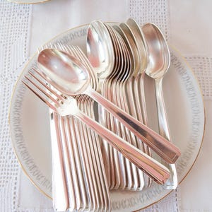 May include: A collection of silver-colored flatware, including spoons and forks, arranged on a white plate with a gold rim. The plate is set on a white, patterned tablecloth. A matching teacup is visible in the background.