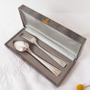 Puede incluir: Un juego de cuchara y tenedor plateados dentro de una caja gris texturizada con un cierre dorado. La cubertería se muestra sobre una tela gris similar al satén. La caja está abierta, revelando los utensilios.