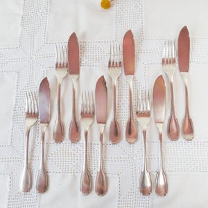 May include: A collection of vintage silver cutlery, including forks and butter knives, is arranged on a white, textured tablecloth. The silverware has a silver finish. A sprig of yellow flowers adds a touch of colour to the scene.