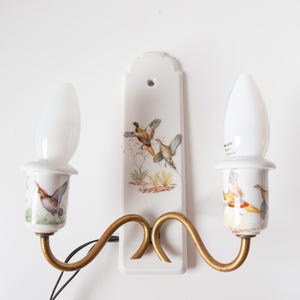 Op de afbeelding: Een vintage wandlamp met twee armaturen. De lamp heeft een witte keramische achterplaat met een geschilderd vogelontwerp en goudkleurige metalen armen. De gloeilampen zijn wit en langwerpig.