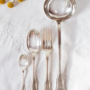 Può includere: Una collezione di posate d'argento antiche, tra cui un mestolo, due cucchiai e una forchetta, disposte su una tovaglia bianca e strutturata. Le posate hanno una finitura argentata riflettente. Un rametto di fiori gialli è in alto a sinistra.