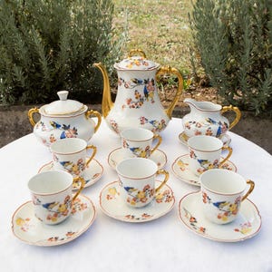 Puede incluir: Un juego de té de porcelana con base blanca y un patrón colorido de pájaros azules y flores naranjas. El conjunto incluye una tetera con tapa, un azucarero con tapa, una jarra de leche y seis tazas con sus platos, todos con asas doradas.