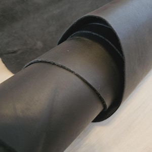 Puede incluir: Primer plano de un rollo de cuero negro, con una superficie lisa y brillante. Los bordes del cuero son visibles, mostrando el grosor y la textura. Un trozo de cuero de ante gris oscuro se encuentra en el fondo.