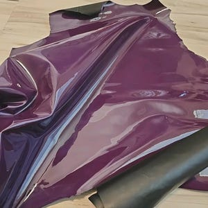 Könnte beinhalten: Ein Bogen aus glänzendem, tiefviolettem Vinylgewebe mit reflektierender Oberfläche. Das Material ist gefaltet und geschichtet, wobei eine schwarze Rückseite sichtbar wird. Der Stoff hat ein glattes, glänzendes Aussehen.