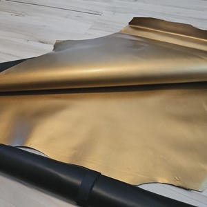 May include: A large sheet of metallic gold leather is partially rolled, revealing its shiny surface. A dark green leather piece is visible beneath the gold sheet. A black, rolled leather piece is in the foreground.