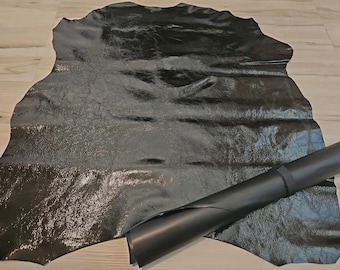 May include: A large, irregularly shaped piece of black leather with a glossy finish. A rolled-up section of the same leather is positioned to the right. The leather is lying on a light-colored wooden surface.