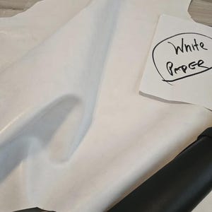 May include: A large piece of white leather with a smooth texture, alongside a rolled-up piece of black leather. A handwritten note on white paper reads "White Paper". The leather appears to be new and unused.
