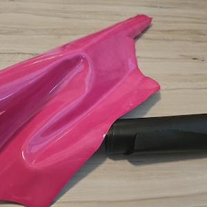 Può includere: Un materiale rosa brillante e lucido è parzialmente arrotolato accanto a un oggetto cilindrico nero. Il materiale rosa ha una superficie liscia e riflettente e l'oggetto nero sembra essere fatto di un materiale opaco.