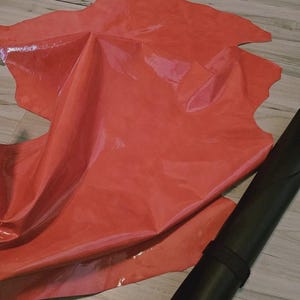May include: A sheet of glossy, red leather with irregular edges is spread out, reflecting light. A rolled-up, black leather piece with a securing strap is positioned nearby. The leather appears to be new and unused.