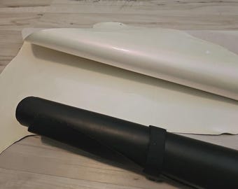 May include: Cream-colored leather hides with a pearlescent sheen are partially rolled and laid out. A rolled black leather mat is secured with a black strap. The items are on a light wood-grain surface.