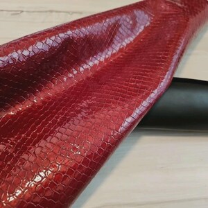 May include: Close-up of a piece of red snakeskin leather with a glossy finish. The leather has a textured, scale-like pattern. A black leather object is partially visible in the background.