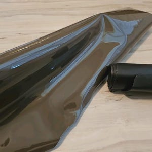May include: A roll of dark gray, shiny, reflective material, possibly vinyl or film, is partially unrolled on a light wood surface. A rolled section of the same material is visible to the right. The material appears to be new and unused.
