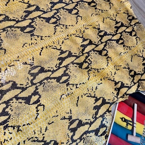 May include: A large piece of yellow leather featuring a black snakeskin pattern. The texture is detailed with small, overlapping scales. In the corner, a small colour swatch card displays various leather samples alongside a metal tool and mallet.