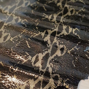 Puede incluir: Primer plano de un patrón de piel de serpiente negra y dorada. El cuero tiene un acabado brillante, con un patrón repetido de escamas doradas y negras. La textura es detallada y el efecto general es lujoso.