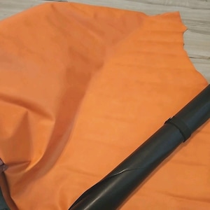 May include: An orange leather hide is spread out, with a rolled-up black leather piece in the foreground. The orange leather has a smooth texture and appears to be a large piece. The black leather is rolled and secured with a black strap.