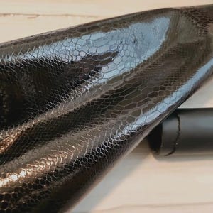Könnte beinhalten: Schwarzes Kunstleder mit Reptilienschuppenmuster. Das Material ist gefaltet, wodurch ein Blick auf die Unterseite gewährt wird. Im Hintergrund ist ein schwarzer zylindrischer Gegenstand zu sehen.