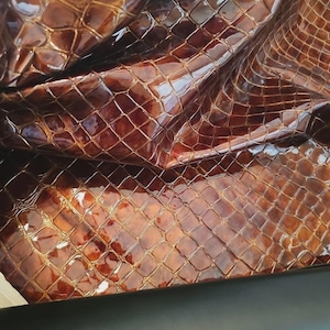 Può includere: Primo piano di un motivo a pelle di serpente marrone lucido. La trama presenta un design a griglia con una base marrone scuro e riflessi marroni più chiari, creando una superficie riflettente. Un materiale nero e liscio è visibile in basso.