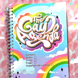 Puede incluir: Un cuaderno con espiral con una cubierta colorida que presenta el texto "The Gay Agenda" en una fuente estilizada de arcoíris. El diseño incluye arcoíris, nubes y estrellas. El cuaderno enumera "Glitter", "Crime", "Frogs" e "Inappropriate content".
