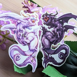 Peut inclure: Deux autocollants colorés représentant un dragon violet et noir et une licorne rose et blanche. Le dragon a des yeux rouges et une grande envergure d'ailes. La licorne a une grande corne et une queue bouclée.