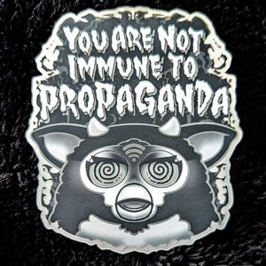 Può includere: Adesivo bianco e nero con un mostro peloso di cartone animato con occhi vorticosi. Il testo "You are not immune to propaganda!" è scritto in bianco, con un carattere irregolare attorno al mostro.
