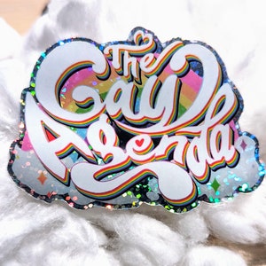 May include: A rainbow-coloured sticker with the text "The Gay Agenda" in white, with a glittery finish.