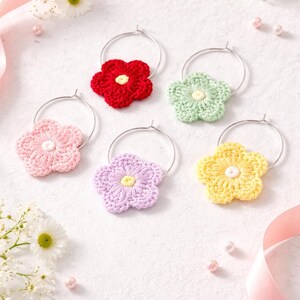 May include: A collection of six colorful, crocheted flower wine charms. Each charm features a different color: red, green, pink, purple, and yellow. The flowers are attached to silver-colored metal hoops. The background includes daisies, pink ribbon, and small pink beads.