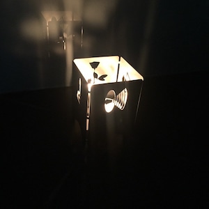 Könnte beinhalten: Ein quadratischer Kerzenhalter aus Metall mit ausgeschnittenen Designs. Der Kerzenhalter ist angezündet und strahlt ein warmes Licht aus.