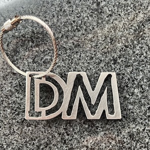 Può includere: Un portachiavi argentato con un ciondolo in metallo che presenta le lettere "DM" in un font stilizzato. Il portachiavi ha un anello in cavo argentato.