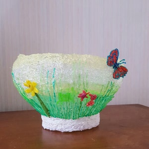 May include: A white ceramic bowl with a green and yellow floral design and a red butterfly. The bowl is decorated with 3D lines that resemble grass and flowers.