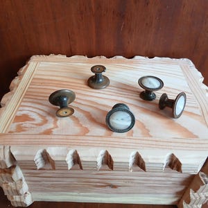 Op de afbeelding: Een houten kist met decoratief gesneden randen en een lichte houten afwerking. De kist is bedekt met verschillende antieke ladeknopen in brons en wit. De knoppen hebben verschillende vormen en ontwerpen.