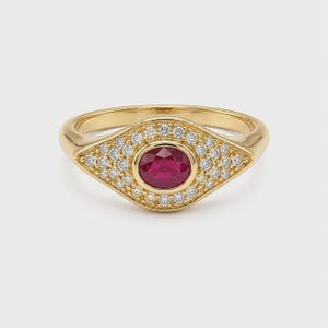 Puede incluir: Un anillo de oro con un rubí rojo intenso de talla ovalada en el centro, rodeado por un halo de pequeños diamantes blancos redondos y claros engastados en un marco con forma de ojo. La banda está hecha de oro amarillo pulido y brillante.