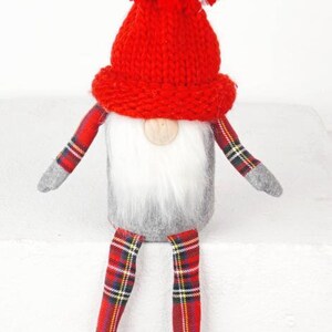 Peut inclure: Une poupée gnome festive avec un bonnet rouge tricoté et un pompon. Le gnome a un corps gris, une barbe blanche et des bras et des jambes à carreaux. La poupée est un objet décoratif.