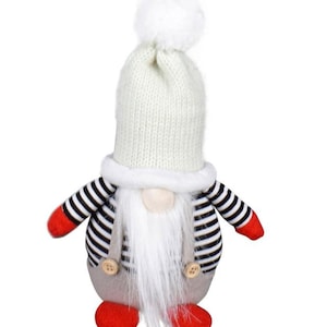 Peut inclure: Figurine de gnome décoratif avec un bonnet en tricot blanc surmonté d'un pompon blanc. Le gnome a une longue barbe blanche, une chemise rayée, un corps gris, des chaussures rouges et de petits boutons en bois.