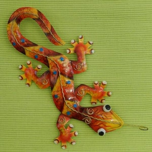 Op de afbeelding: Een kleurrijk metalen wandkunstwerk in de vorm van een hagedis. De hagedis heeft een levendig ontwerp met oranje, gele en rode tinten, geaccentueerd met blauwe stippen. De staart van de hagedis krult en de voeten hebben een stervorm.