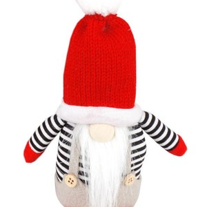 Peut inclure: Un gnome de Noël avec un bonnet rouge tricoté et un pompon blanc. Le gnome a une longue barbe blanche, un nez en bois et porte une chemise rayée noire et blanche avec un pantalon gris. Des chaussures rouges complètent la décoration.