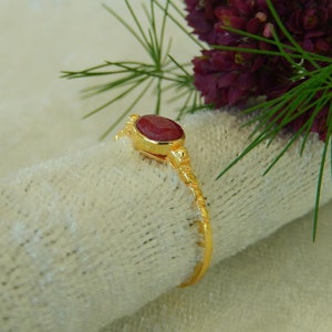 Op de afbeelding: Een gouden ring met een rode ovale edelsteen in een bezel-setting. De ringband is dun en heeft een getextureerd ontwerp.