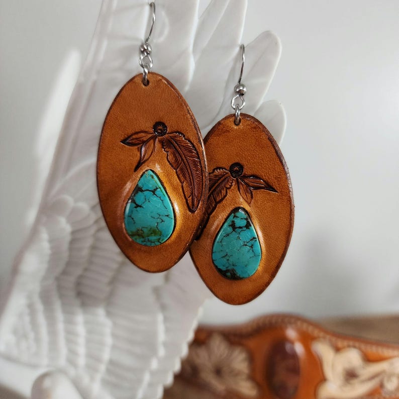 May include: A pair of oval-shaped leather earrings with turquoise stones and a feather design. The earrings are brown with a light brown trim.