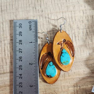 May include: A pair of leather earrings with turquoise stones and a feather design. The earrings are approximately 2 inches long.