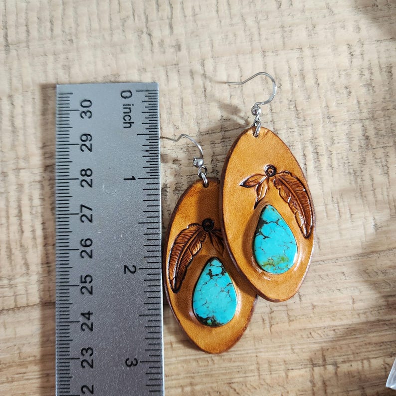 May include: A pair of leather earrings with turquoise stones and a feather design. The earrings are approximately 2 inches long.