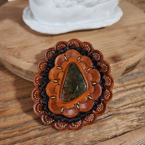 May include: A leather pendant with a green agate stone set in the center. The pendant is decorated with black and yellow stitching in a floral pattern.