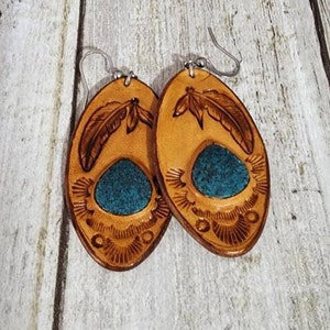 May include: A pair of oval-shaped leather earrings with a turquoise inlay and a carved feather design. The earrings are brown with a light brown border.
