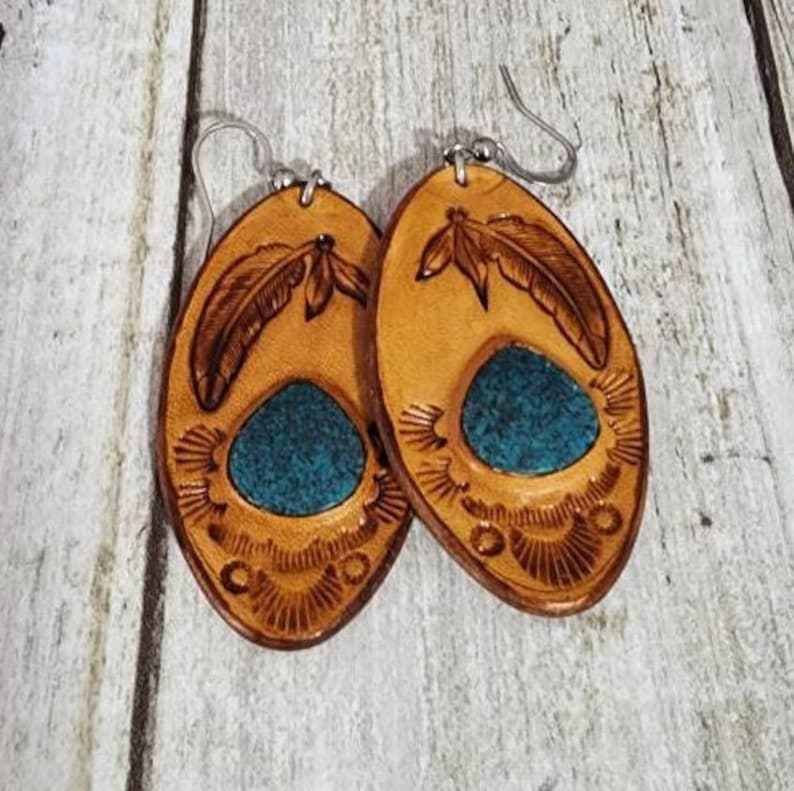 May include: A pair of oval-shaped leather earrings with a turquoise inlay and a carved feather design. The earrings are brown with a light brown border.