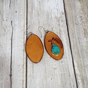 May include: A pair of oval-shaped leather earrings with a turquoise stone inlay. One earring has a dragonfly design carved into the leather.