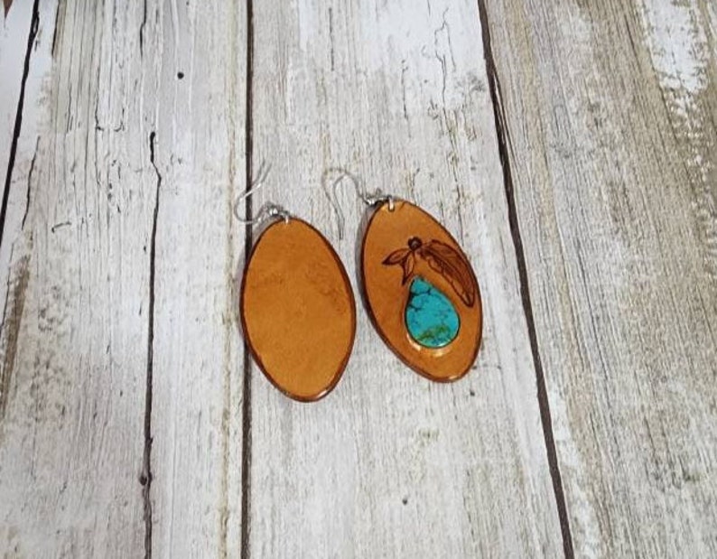 May include: A pair of oval-shaped leather earrings with a turquoise stone inlay. One earring has a dragonfly design carved into the leather.